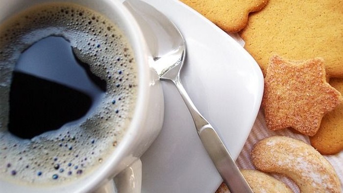 Eine Tasse Kaffee mit Latte Art steht neben festlichen sternförmigen Keksen auf einer Untertasse mit einem Löffel.
