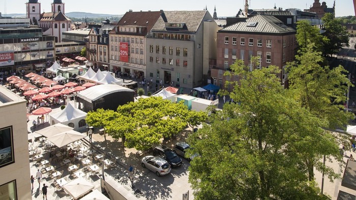 Ein belebter europäischer Marktplatz, gefüllt mit Marktständen, gesäumt von historischen Gebäuden und Grünflächen.