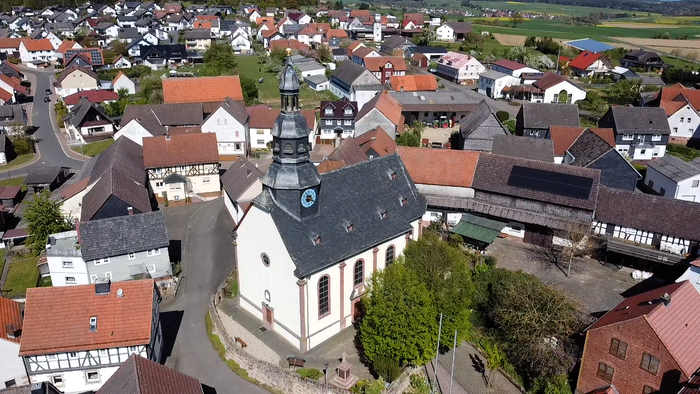 Luftaufnahme eines malerischen Dorfes mit einer markanten Kirche, umgeben von bunten Häusern und üppigem Grün.