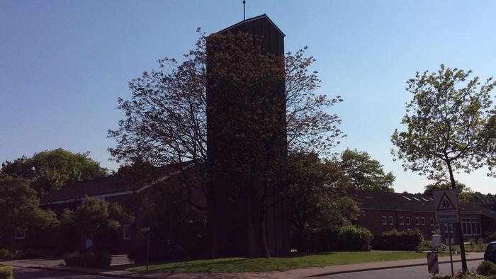 Kirchturm mit Kreuz auf dem Dach, umgeben von Bäumen und Gebäuden, klarer blauer Himmel