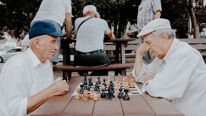 Zwei ältere Männer, die sich intensiv auf ein Schachspiel im Freien konzentrieren, während andere im Hintergrund zuschauen.