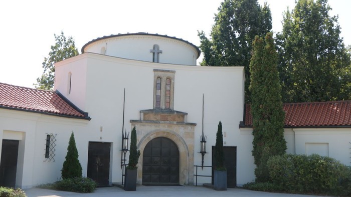 Das Bild zeigt ein weißes Gebäude mit Kuppel und Kreuz, umgeben von Bäumen und Büschen.