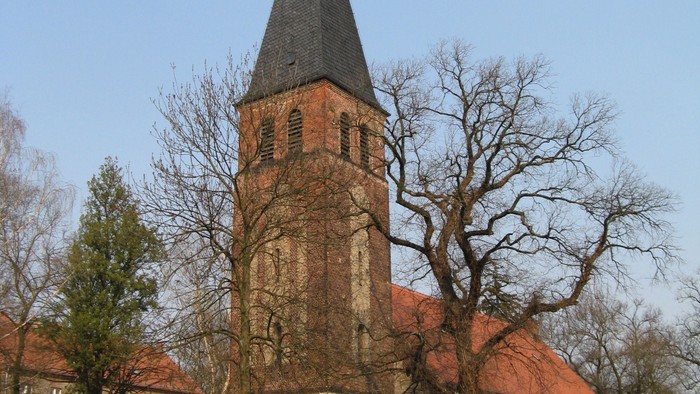 Historische Steinkirche mit hohem Backsteinturm und steilem Satteldach, umgeben von kahlen Bäumen.