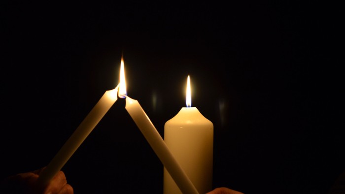 Zwei brennende Kerzen, die von Händen in einer dunklen Umgebung gehalten werden und mit ihren Flammen eine dreieckige Form bilden.