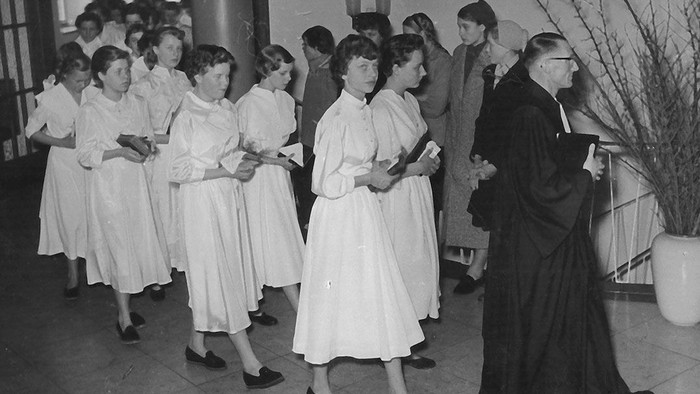 Gruppe von Frauen in weißen Kleidern, die während einer formellen religiösen Zeremonie mit einem Geistlichen Bücher halten.