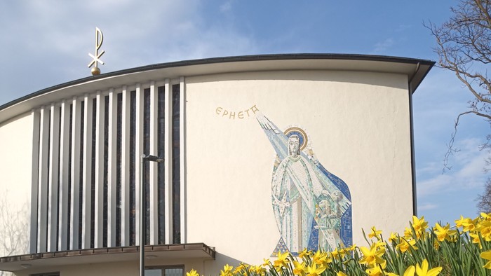 Moderne Kirche mit Wandgemälde eines Heiligen, umgeben von leuchtend gelben Osterglocken im Frühling.