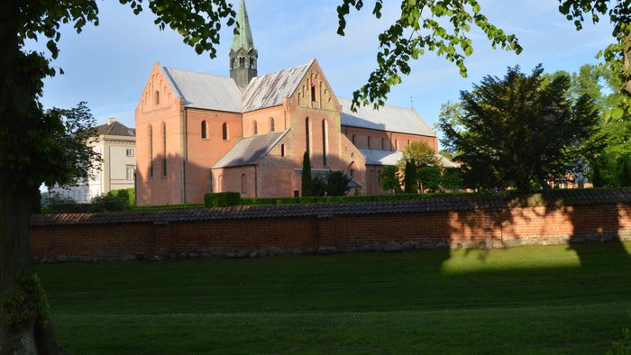 Historisk murstenskirke med et højt spir omgivet af frodig grønne områder og en stensmur.