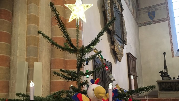 Et festligt juletræ i en kirke, pyntet med lys, stjerner og farverige dekorationer, herunder et tøjdyr.