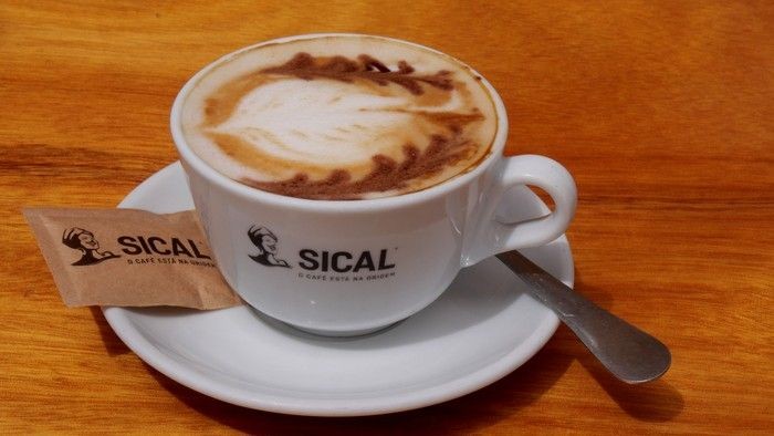 Das Foto zeigt eine gefüllte Cappuccinotasse mit Verzierung im Schaum und einem Logo, sowohl an der Tasse aals auch beim Zucker