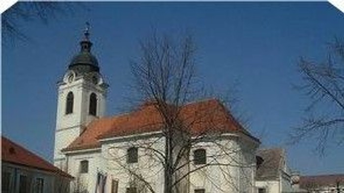 Ein weißes Gebäude mit roter Ziegeldach und hoher Kirchturm vor blauem Himmel.