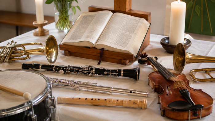 Mehrere Musikinstrumente – darunter Trompete, Klarinette, Quer- und Blockflöte, Geige, Posaune und Trommel – liegen auf einem weiß gedeckten Altar um eine aufgeschlagene Bibel, flankiert von zwei brennenden Kerzen und einem Holzkreuz.