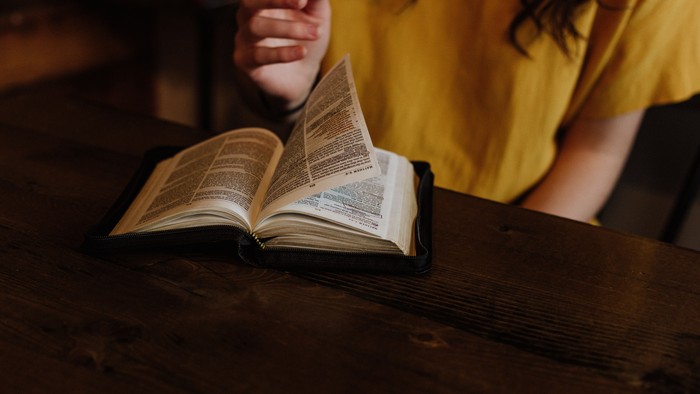 Eine Person in einem gelben Hemd liest ein aufgeschlagenes Buch an einem hölzernen Tisch.