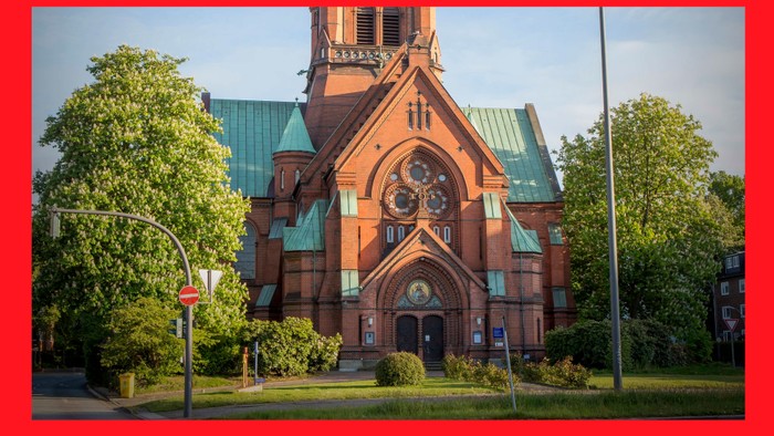 Das Bild zeigt eine rote Kirche mit grünen Dächern und einem Uhrturm, umgeben von Bäumen und Gras.