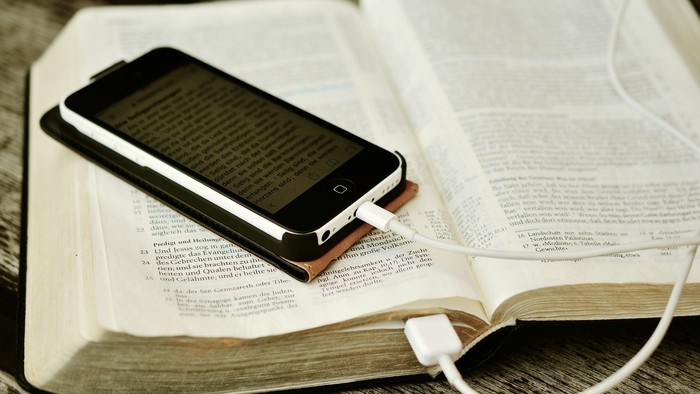 Ein Smartphone liegt auf einem aufgeschlagenen Buch, an das ein Ladekabel angeschlossen ist.