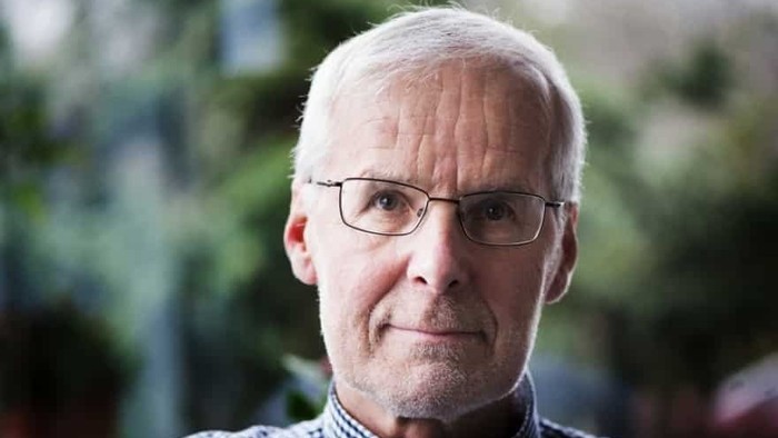 En mand med briller og skaldet hoved iført en ternet skjorte foran en sløret udendørs baggrund.