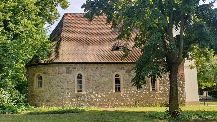 Altes Steinhaus mit spitzem Dach und Fensterbögen, umgeben von Bäumen und Gras.