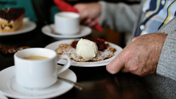 Eine Person hält einen Teller mit Kuchen und Eiscreme, daneben stehen Tassen mit Kaffee auf dem Tisch.