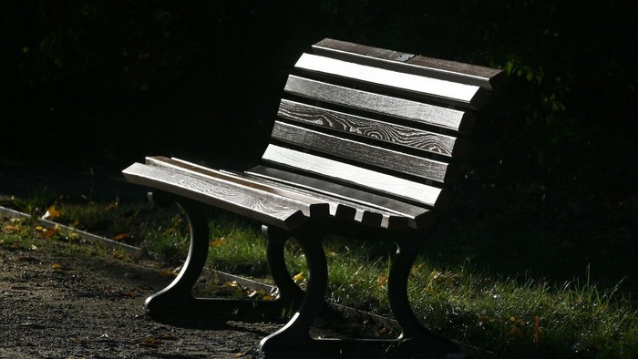 Eine einsame Holzbank wirft Schatten auf einen schwach beleuchteten Weg, umgeben von Grün.