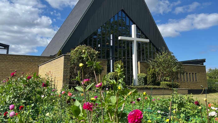 Moderne kirke med et højt kors mod en klar blå himmel, omgivet af farverige blomster.