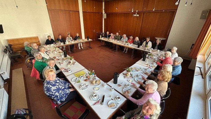 Eine Gruppe von Menschen sitzt an langen Tischen in einer Speisehalle, unterhält sich und genießt gemeinsam eine Mahlzeit.