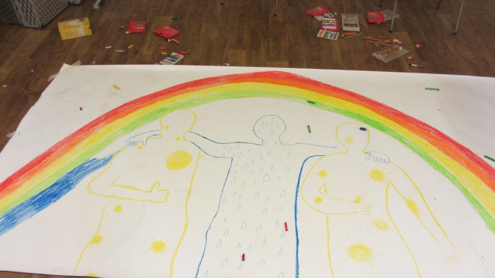 Ein großes Papier mit einem gemalten Regenbogen und zwei Figuren liegt auf dem Boden, umgeben von Malutensilien.