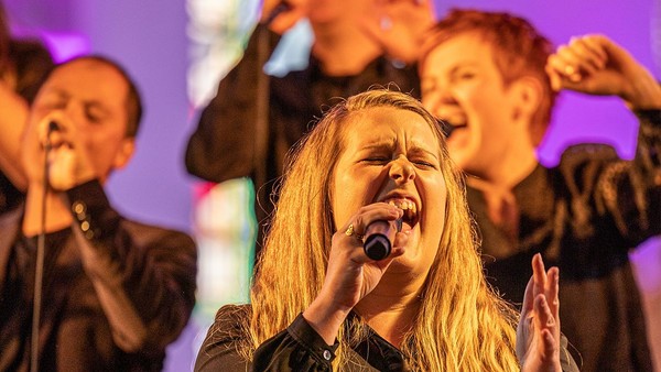 Ein Mädchen singt leidenschaftlich in ein Mikrofon, umgeben von anderen singenden Kindern auf einer Bühne.