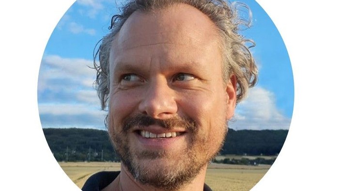 Lächelnder Mann mit lockigem Haar vor einer hellen, offenen Landschaft im Freien.
