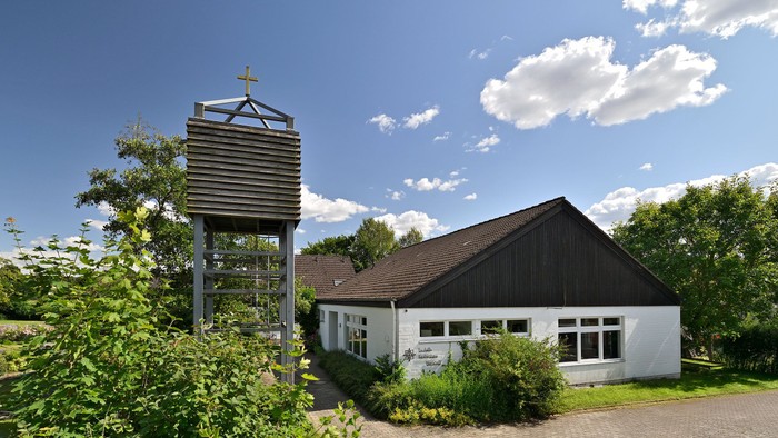 Eine weiße Kapelle mit einem braunen Spitzdach. Der Glockenturm steht links neben der Kapelle. 