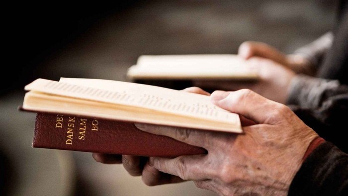 Hænder, der holder en rød dansk bibel åben med tekst på dansk, sandsynligvis under læsning i en gudstjeneste.