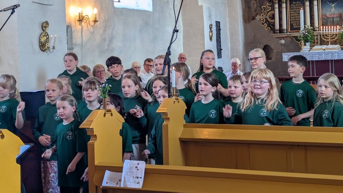 En ungdomskor i grønne uniformer optræder i en kirke og synger med begejstring og mikrofoner.