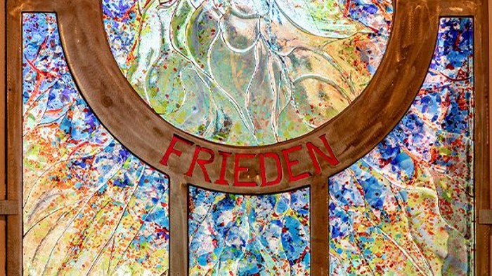 Buntglasfenster mit dem Wort „Frieden“, farbenfrohen abstrakten Mustern und einer zentralen Figur.