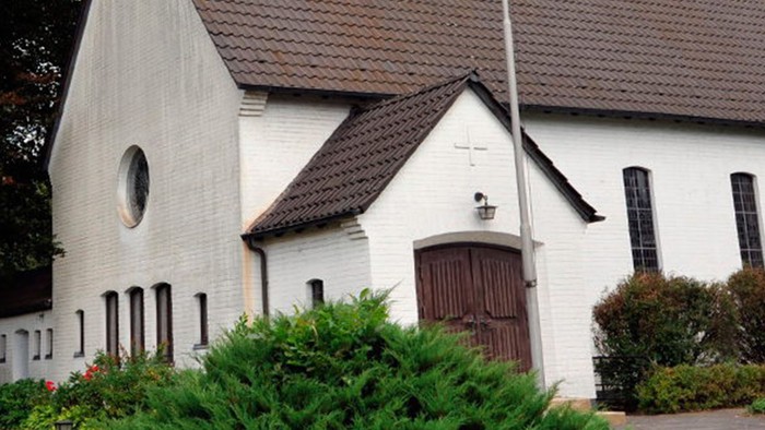 Weiße Kirche mit hohem Turm und braunem Dach, umgeben von Bäumen und Büschen