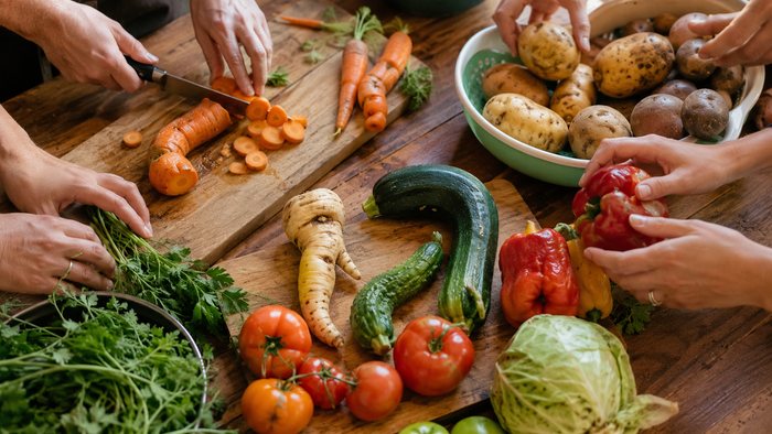 Mehrere Hände bereiten gemeinsam frisches Gemüse auf einem Holztisch zu: Karotten werden geschnitten, Kartoffeln sortiert und Paprika gehalten; dazwischen liegen Tomaten, Gurken, Zucchini, Kohl, Petersilie und weiteres Gemüse.