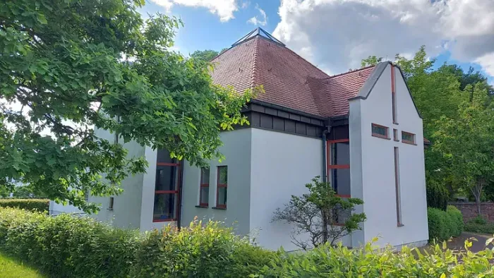 Modernes Haus mit rotem Ziegeldach, umgeben von üppigem Grün unter einem teilweise bewölkten Himmel.