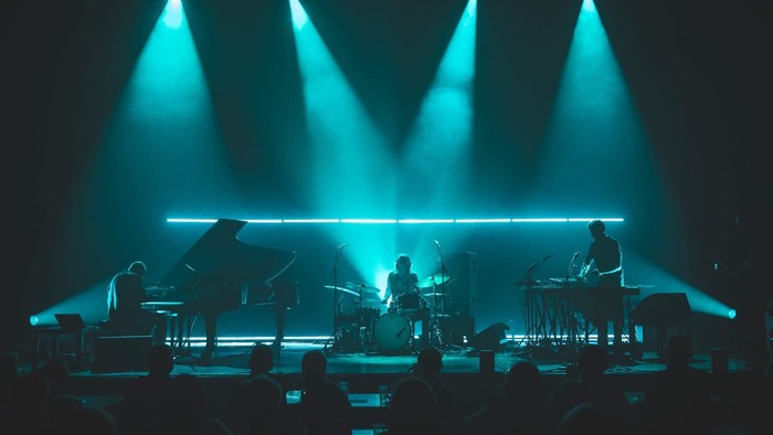 Eine Live-Band tritt unter dramatischer blauer Bühnenbeleuchtung mit einem Pianisten, einem Schlagzeuger und einem DJ auf.