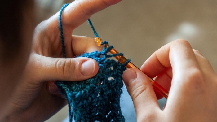 Strickende Hände mit blauem Garn und orangen Stricknadeln.
