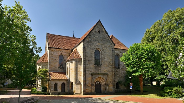 Historische Steinkirche mit steilem, rotziegelgedecktem Dach und bogenförmigem Eingang, umgeben von üppigem Grün.