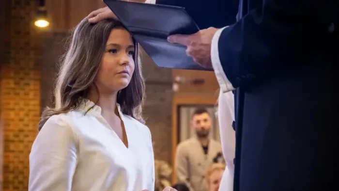 En kvinde i hvidt tøj modtager et dokument fra en mand ved en formel begivenhed eller ceremoni.