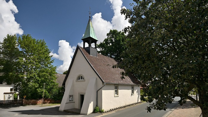 Idyllische Kirche mit steilem Dach und grünem Turm, eingebettet zwischen üppigen grünen Bäumen.