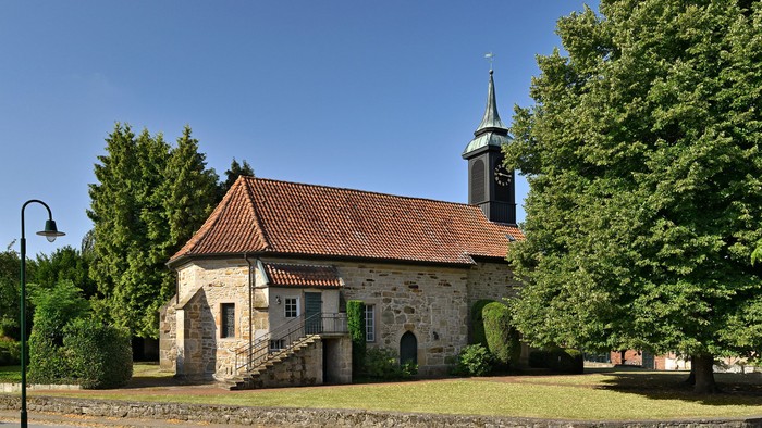 Idyllische Steinkirche mit steilem Ziegeldach und hohem Turm, umgeben von üppigem Grün.