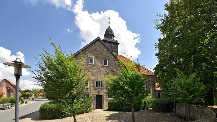 Historische Steinkirche mit Glockenturm, umgeben von üppigem Grün und einem Kopfsteinpflasterweg.