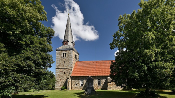 Historische Steinkirche mit hohem Turm, eingerahmt von üppigen grünen Bäumen unter einem klaren blauen Himmel.