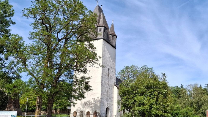 Historischer Steinkirchturm, umgeben von üppigem Grün unter einem klaren blauen Himmel.
