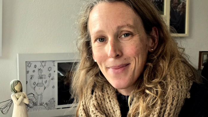 Frau hält Holzfigur, lächelt, trägt einen Schal, mit gerahmten Bildern an der Wand.