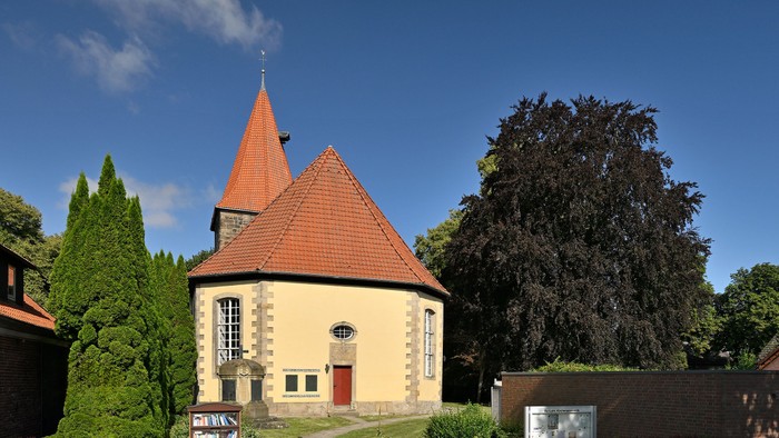 Idyllische Kirche mit rotem Ziegeldach und spitzem Turm, umgeben von üppigem Grün unter klarem blauem Himmel.