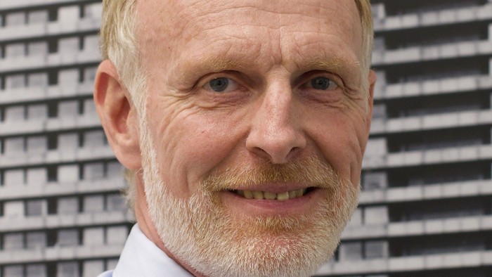 Mand med smil iført en lyseblå skjorte og et mønstret slips foran en geometrisk mønstret baggrund.