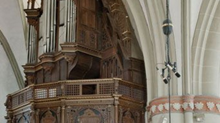 Komplexe hölzerne Kirchenorgel mit verzierten Pfeifen und dekorativem Kronleuchter in historischem architektonischem Ambiente.