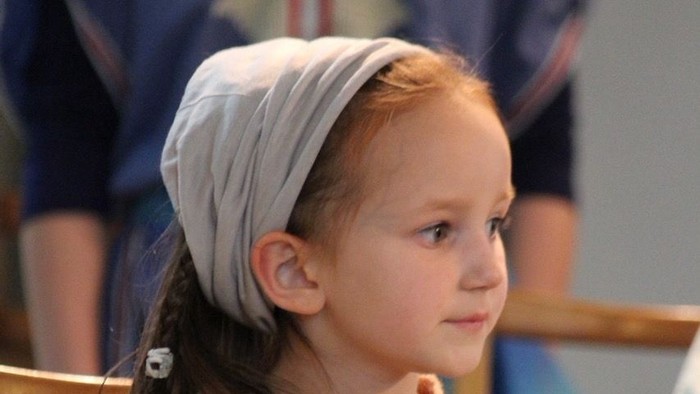 Junge Frau in historischem Gewand mit Kopftuch und Strickgürtel, aufmerksam im Innenraum sitzend.