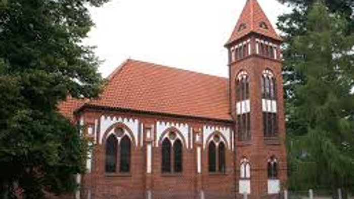 Das Bild zeigt eine rote Backsteinkirche mit hohem Turm und spitzen Fenstern, umgeben von Bäumen und einem Zaun.