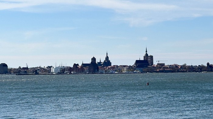 Ein ruhiger Blick auf eine Uferpromenade mit der Skyline einer fernen Stadt unter klarem blauem Himmel und einem Drachen, der darüber schwebt.
